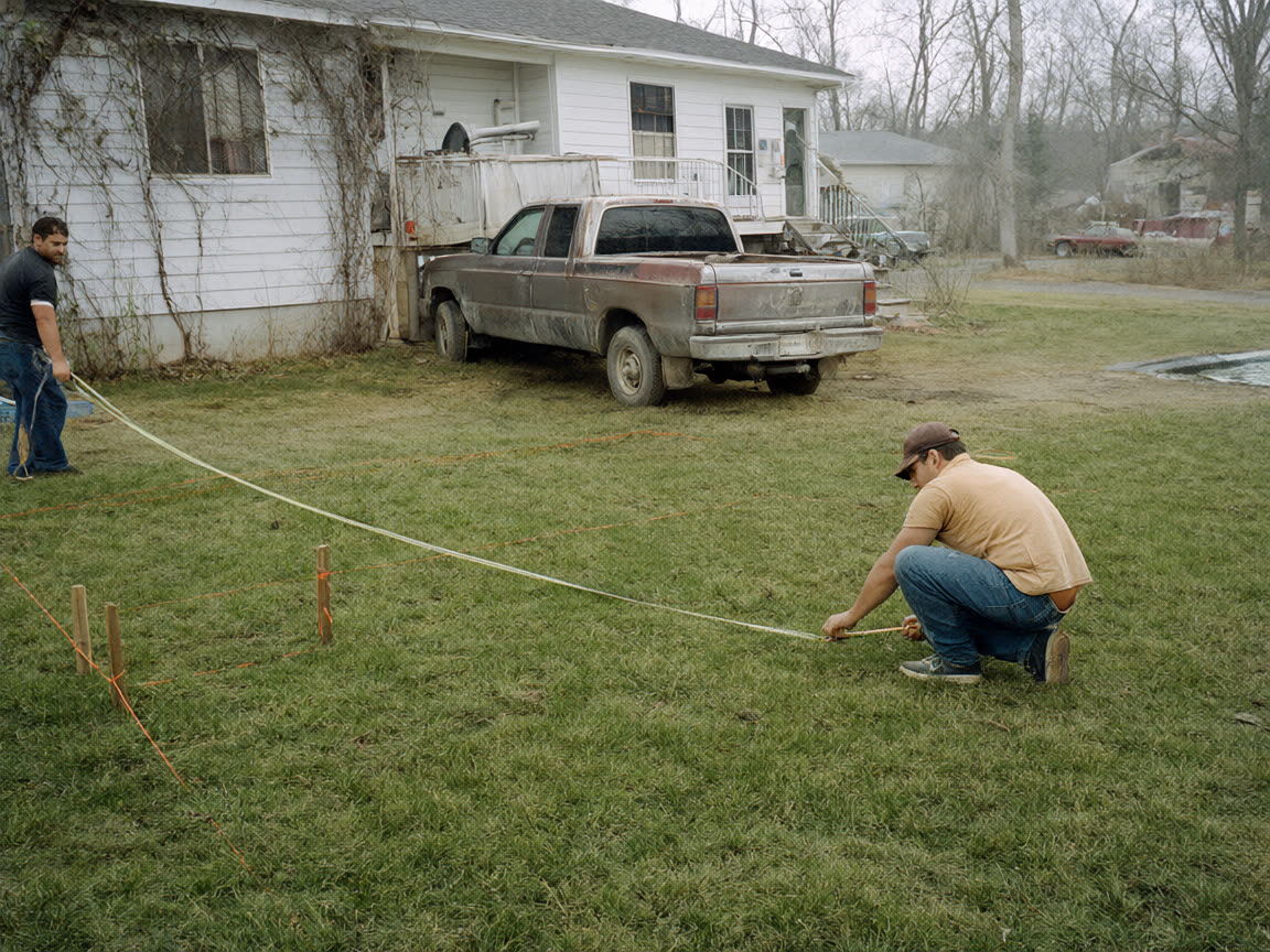 Two men stretching a tape measure across a lawn between wooden stakes and orange string, a muddy pickup parked by the house